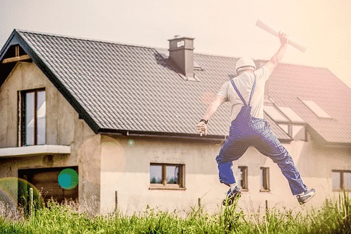 A construction worker in overalls and a hardhat jumping excitedly in front of a house.
